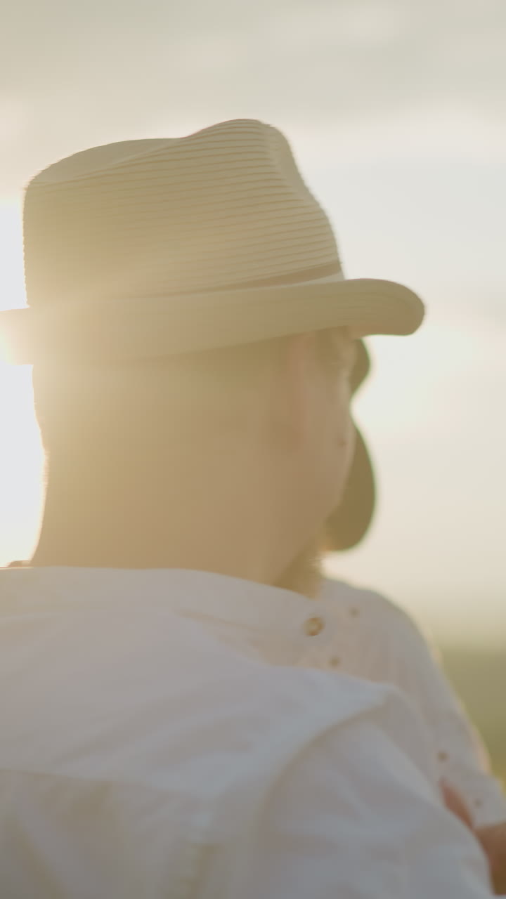 A close-up of a joyful husband and wife dancing together at sunset, both dressed in white shirts and hats. The warm, golden light highlights their happy expressions