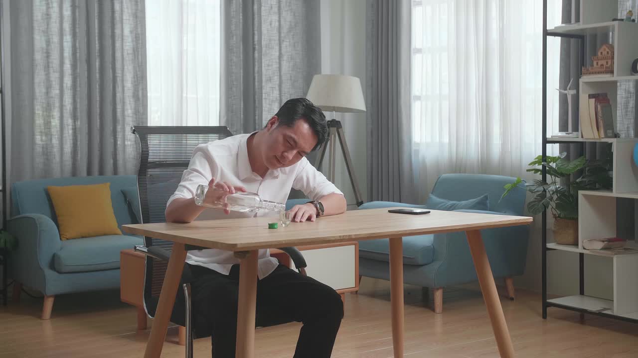 술에 취한 아시아인 남자가 테이블 위에 스마트폰을 들고 글라스에 보드카를 부어서 집에서 술을 마시기 전에 카메라에 환호합니다.