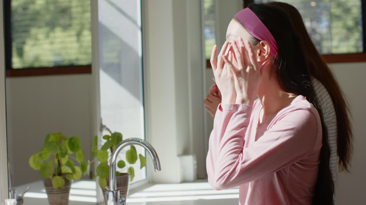 Applying skincare, asian teenage girl in pink headband standing by kitchen sink with plant