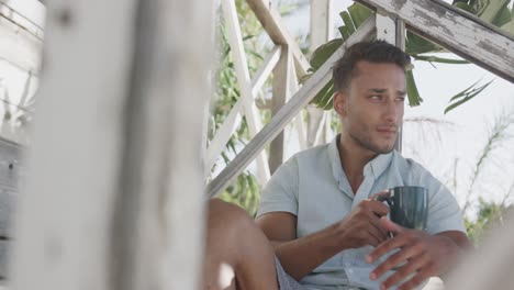 Happy-biracial-man-drinking-coffee-sitting-on-porch-of-beach-house,-in-slow-motion