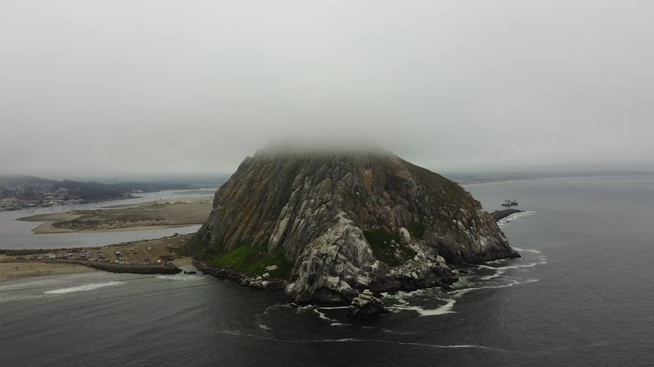 Counterclockwise drone shot of Morro Rock peaking through the clouds on a foggy day on the California coast. Waves crash onto the nearby beach as a ship makes its way to sea from Morro Bay. 4k aerial.