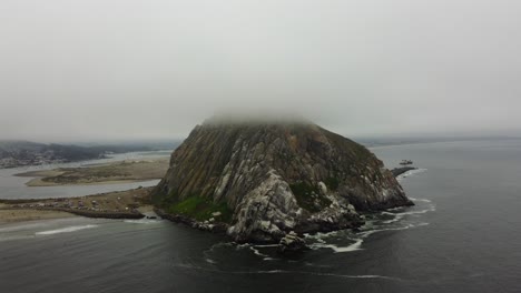 counterclockwise drone shot of morro rock peaking through the clouds on a foggy day on the california coast. waves crash onto the nearby beach as a ship makes its way to sea from morro bay. 4k aerial.