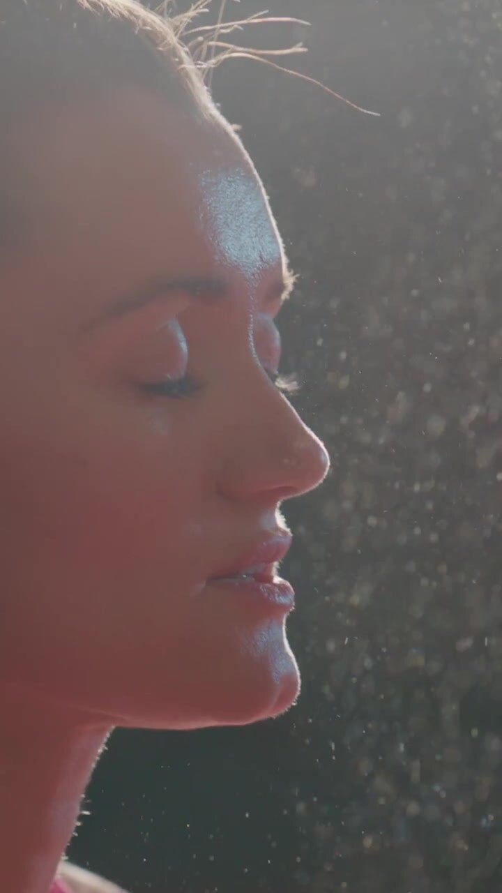 Woman's face, illuminated by warm sunlight. Dust particles float around her in soft light. She appears calm and focused, eyes gently closed. Her hair glows as sunlight catches it