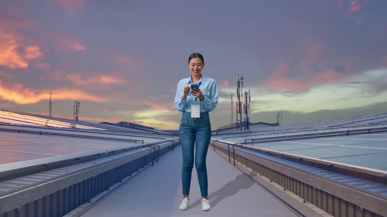 Figura intera di una donna asiatica con il suo smartphone, che ispeziona pannelli solari industriali sul tetto, lavorando ininterrottamente.