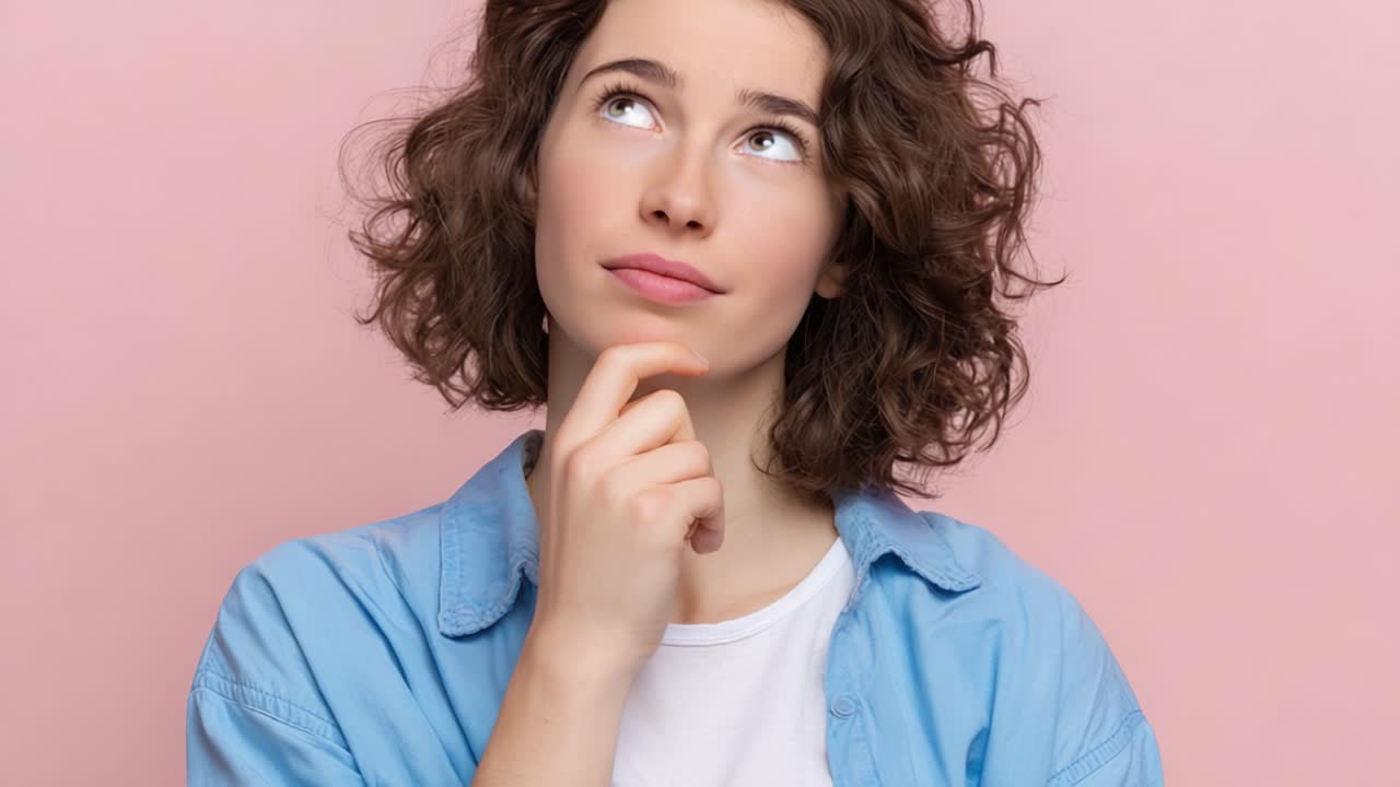 Een bedachtzame jonge vrouw die diep in gedachten is met een nieuwsgierige uitdrukking tegen een zachtroze achtergrond, wat een reflecterend moment van introspectie en verwondering toont.