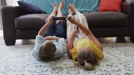 happy caucasian brother and sister at home, lying on floor in living room using tablet together