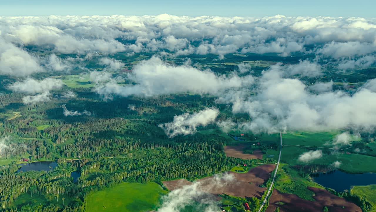 A stunning high-altitude aerial timelapse captures turbulent white clouds moving like a river over the vast green landscape of the Latvian countryside