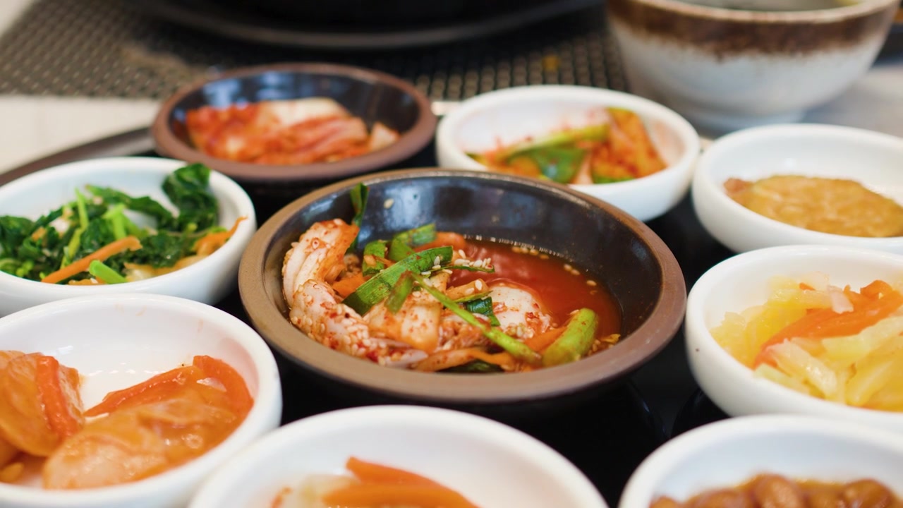 Tampak dekat berbagai lauk Korea dengan seseorang sedang makan semur di latar belakang.