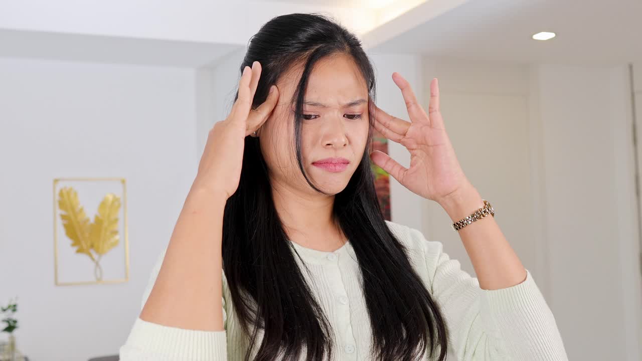 A woman appears distressed, holding her head in a well-lit room with minimal decor