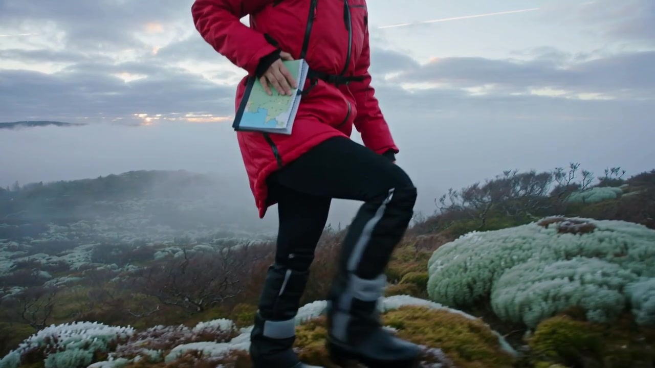 Stepping from moss mound, hiker striding up coastal slope to ridge, wearing red jacket holding map