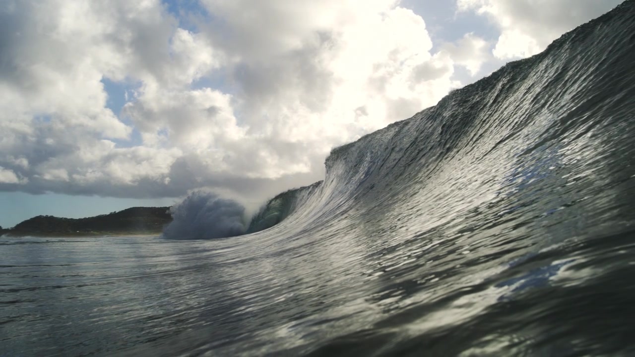 Steep ocean wave breaks into white spray beneath bright clouds in detailed slow motion