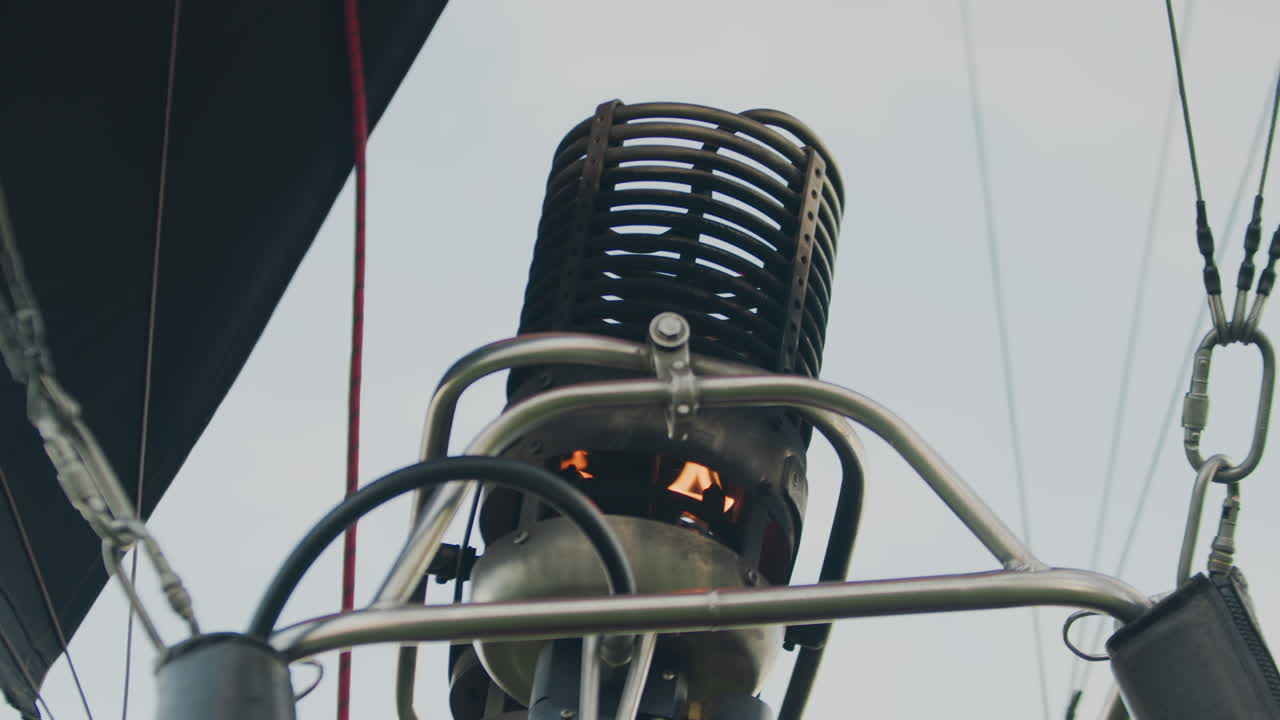 Close up of metal burner on hot air balloon with small visible flame igniting inside cylindrical structure, framed by steel pipes and wires, under bright sky during pre flight setup phase