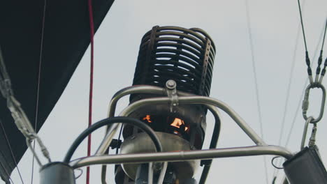 close up of metal burner on hot air balloon with small visible flame igniting inside cylindrical structure, framed by steel pipes and wires, under bright sky during pre flight setup phase