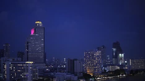 An-aerial-view-of-the-lively-nightlife-along-Chao-Phraya-River-in-Bangkok,-showcasing-the-glistening-city-lights-and-passing-boats