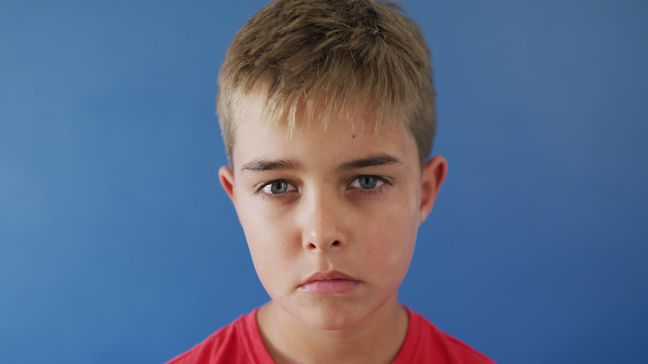 Close up of portrait of sad caucasian boy looking at camera on blue background, slow motion