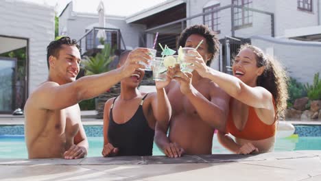 group of diverse male and female friends toasting with drinks and laughing in swimming pool