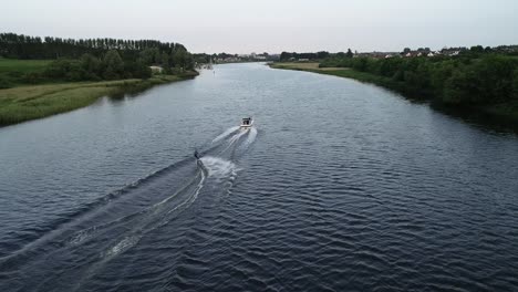 Seguimiento-De-La-Vista-Aérea-De-Un-Esquiador-Acuático-Y-Un-Bote-Remolcador-En-Un-Río-Tranquilo,-Coleraine,-Irlanda-Del-Norte,-Gran-Bretaña,-Disparo-De-Drones-De-60-Fps