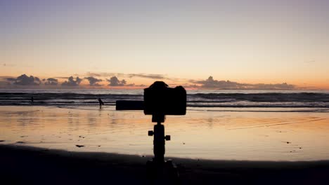 a camera is set up on a tripod to take pictures of the stunning view of the sunset at low tide in laguna beach as surfers surf and the sun peaks out amongst rocks and reflects off the water