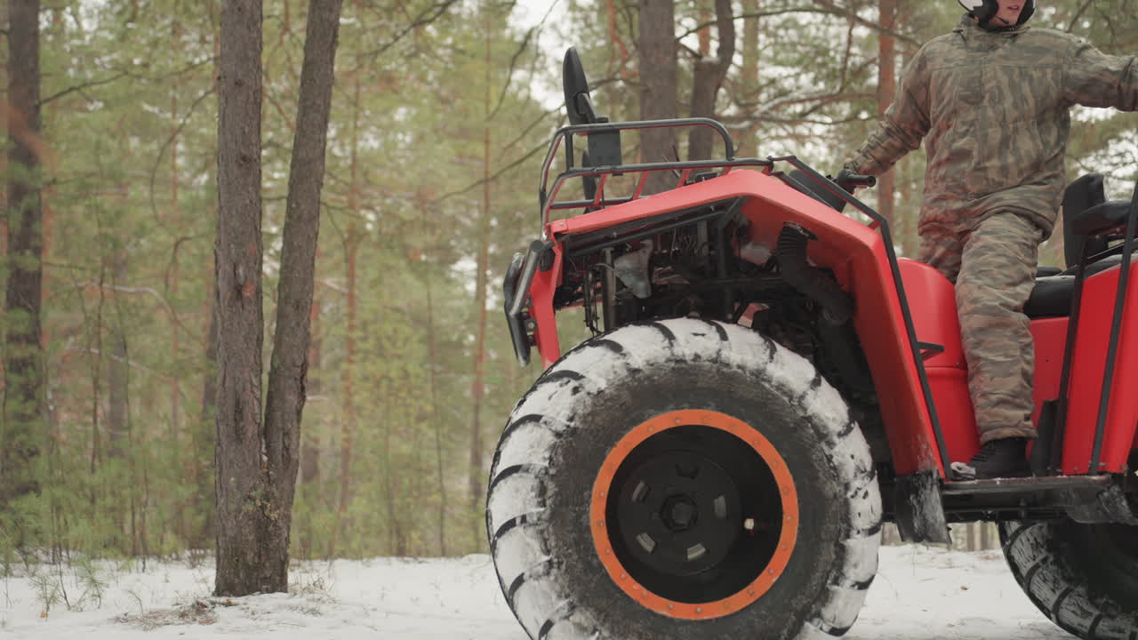 Un pilota di ATV in una foresta innevata ispeziona un quad rosso indossando una tuta mimetica e un casco bianco, con il terreno coperto di neve e pini. Primo piano dei pneumatici tassellati, mentre controlla l'acceleratore e la marcia, preparandosi a salire, in un'atmosfera tranquilla.