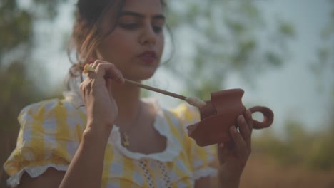 young woman standing in a field holding a stick with glue on it and applying it to a broken clay cup