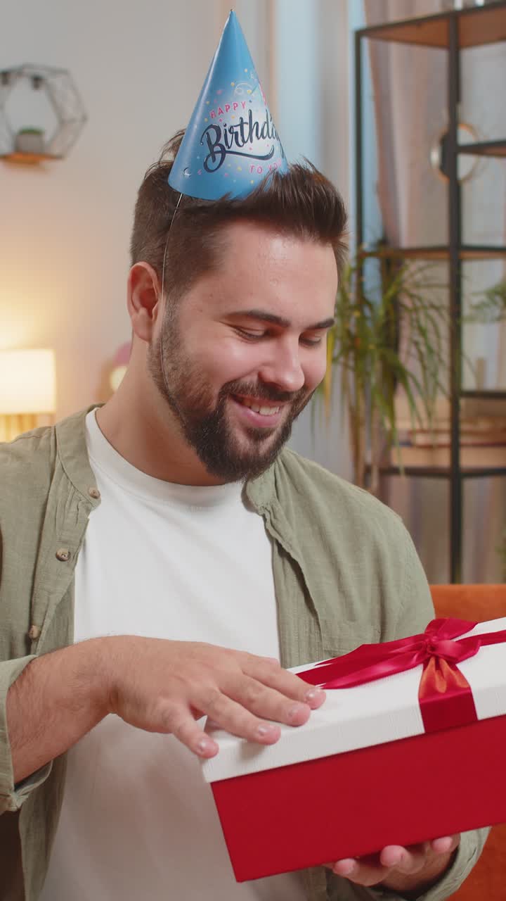 Un giovane felice e sorpreso indossa un cappello di compleanno festivo aprendo una scatola regalo sul divano a casa