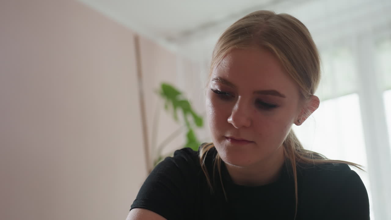 Pretty blonde woman with hair tied back wearing black shirt looking downward in focus, surrounded by softly lit room with blurred plant and window background creating a calm, serene mood