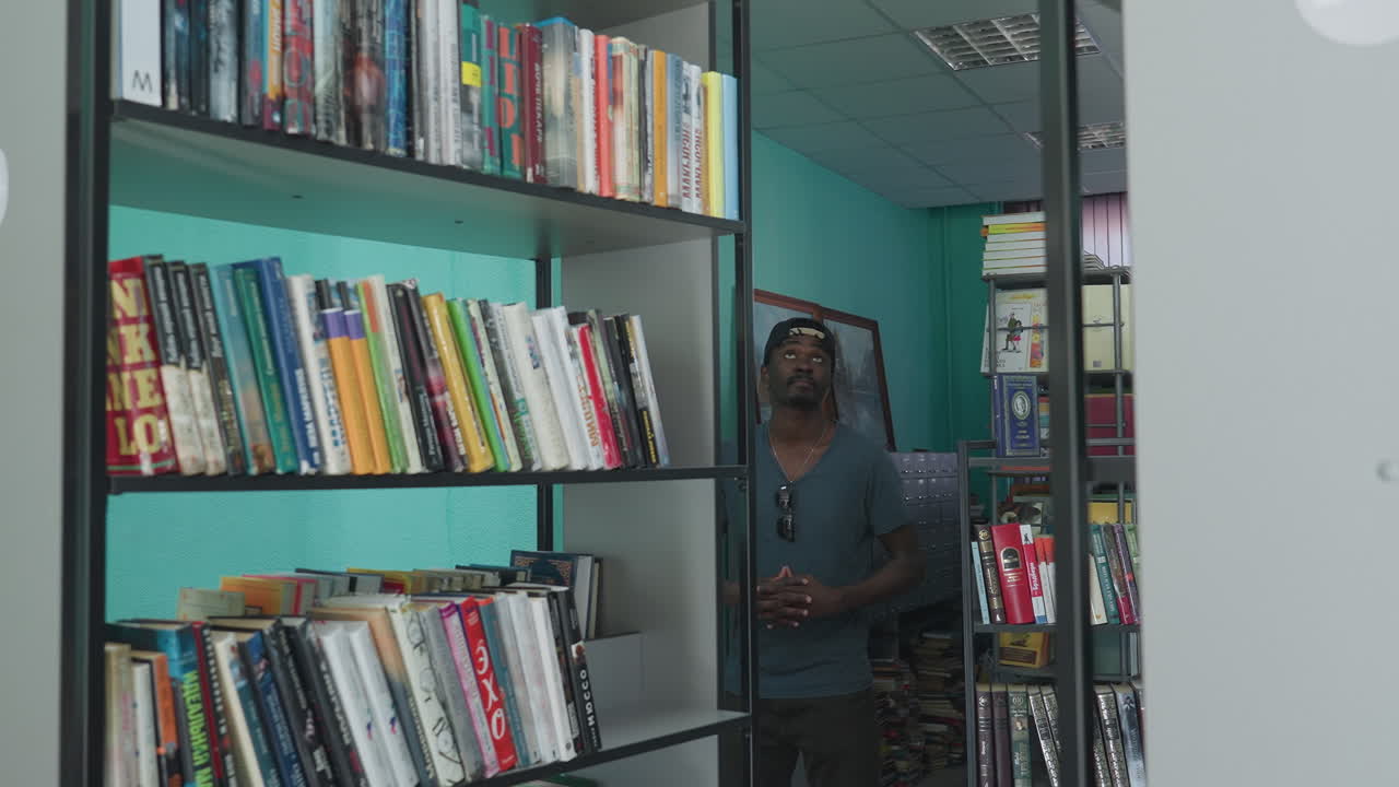 Academician wearing casual shirt and cap stands between library shelves, looking around thoughtfully with hands joined, surrounded by colorful books and vibrant interior in quiet study environment