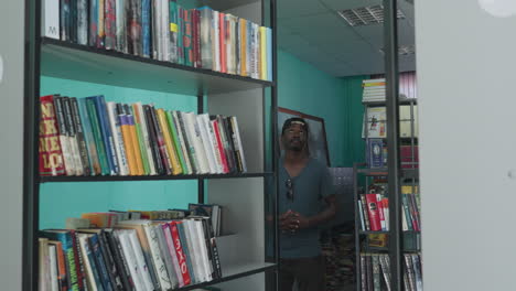 academician wearing casual shirt and cap stands between library shelves, looking around thoughtfully with hands joined, surrounded by colorful books and vibrant interior in quiet study environment