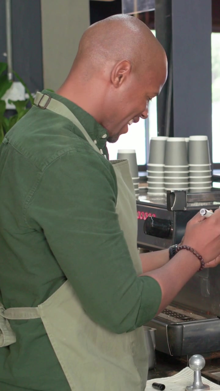 Vertical video: Placing cup barista in apron at counter making coffee steaming milk with pitcher