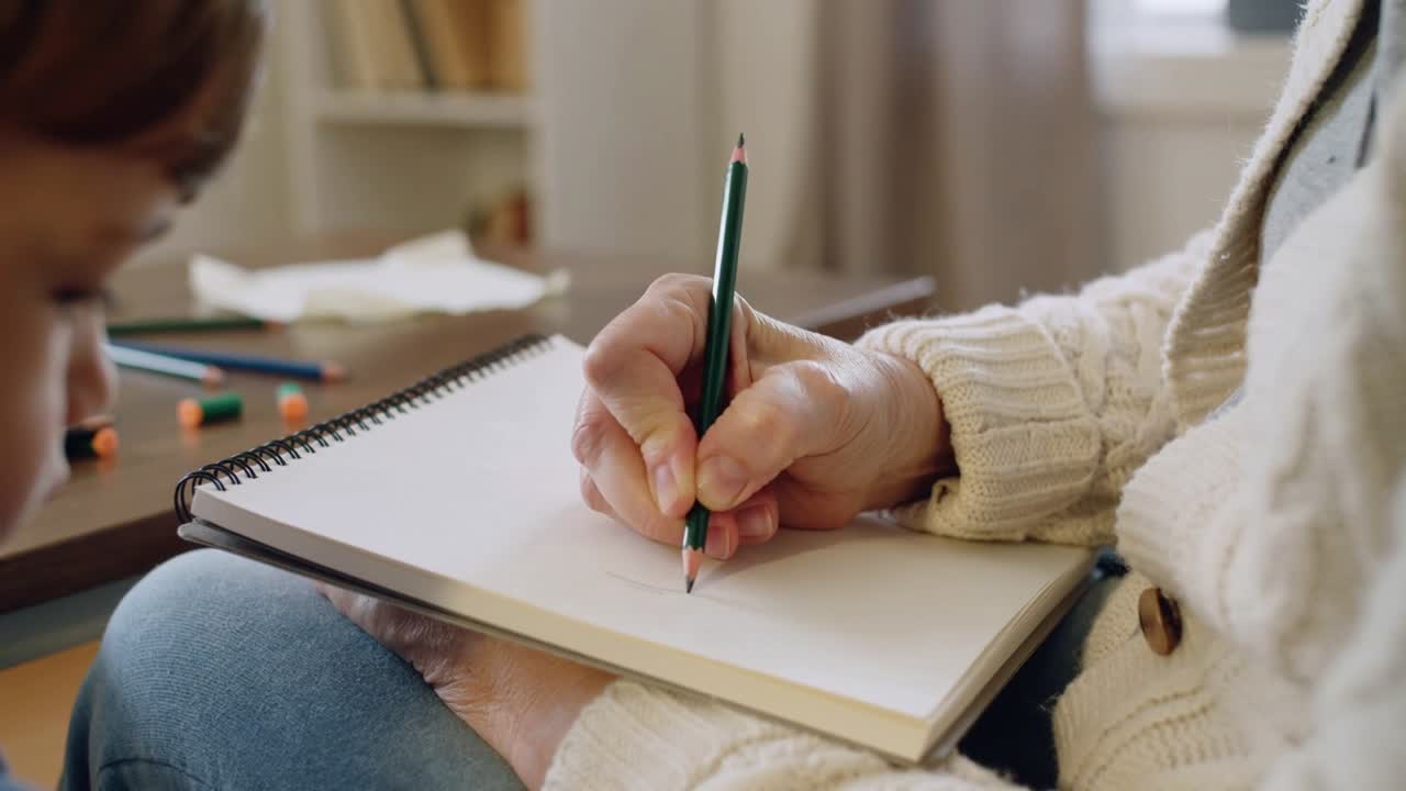 Badante che abbassa una matita verde su un quaderno da disegno appoggiato sul grembo con un bambino che guarda, mentre fa piccoli segni per insegnare l'arte.