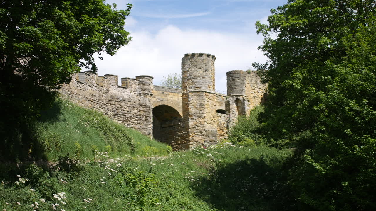 A stone castle gate stands partly shaded by trees in Scarborough, North Yorkshire, England, with a winding path and tall grass leading toward the historic ruins