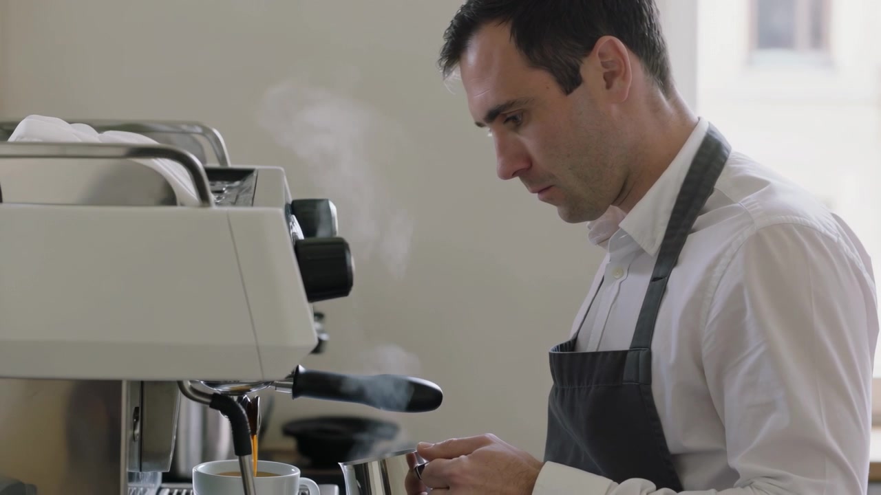 Pulling espresso, wand steaming, barista gray-apron holding pitcher at counter serving, copy space