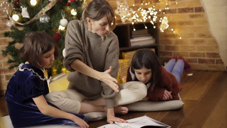 mamá con niños sentados en el suelo leyeron un libro juntos en el fondo guirnaldas de luces navideñas