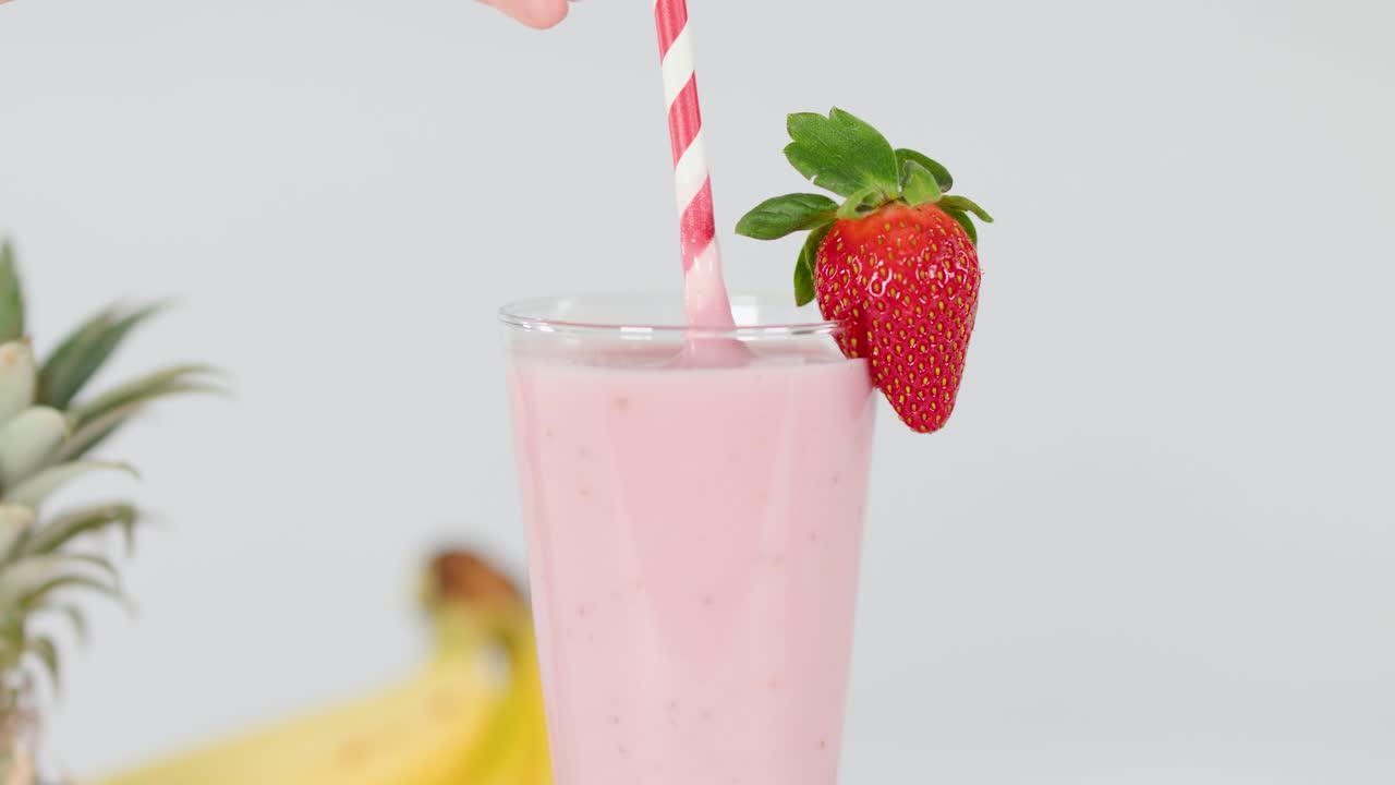 Hand inserts striped straw into creamy pink smoothie with fresh fruit garnish, bright studio lighting