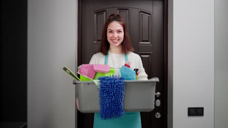 Portrait-of-a-confident-young-brunette-cleaning-lady-in-a-blue-apron-who-holds-in-her-hands-a-gray-basin-with-various-cleaners-and-items-for-cleaning-the-apartment