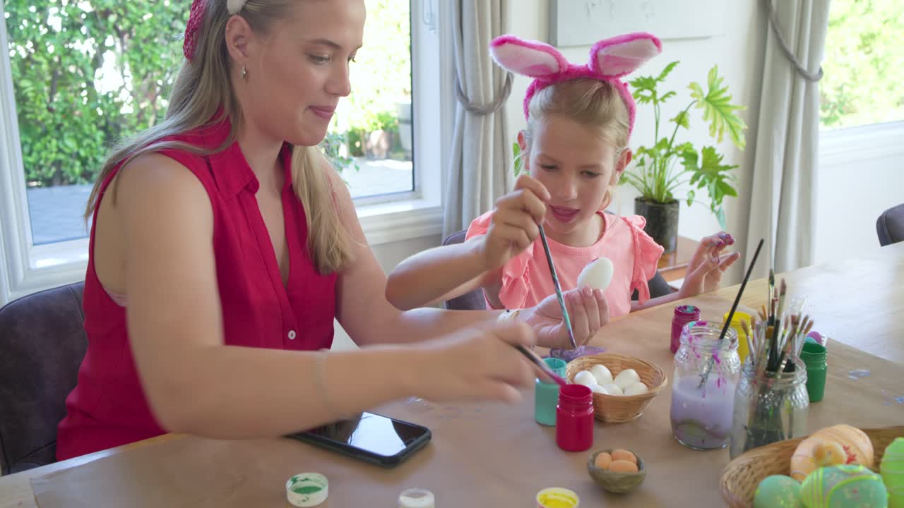 Mother and daughter at table wearing bunny ears daughter selecting brush decorating eggs
