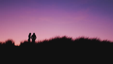 silhouette of couple at beach standing on sand dunes covered in grass against setting sun