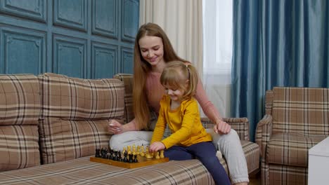 Small-cute-daughter-child-and-young-mother-playing-chess-at-home-sofa,-leisure-hobbies-activities