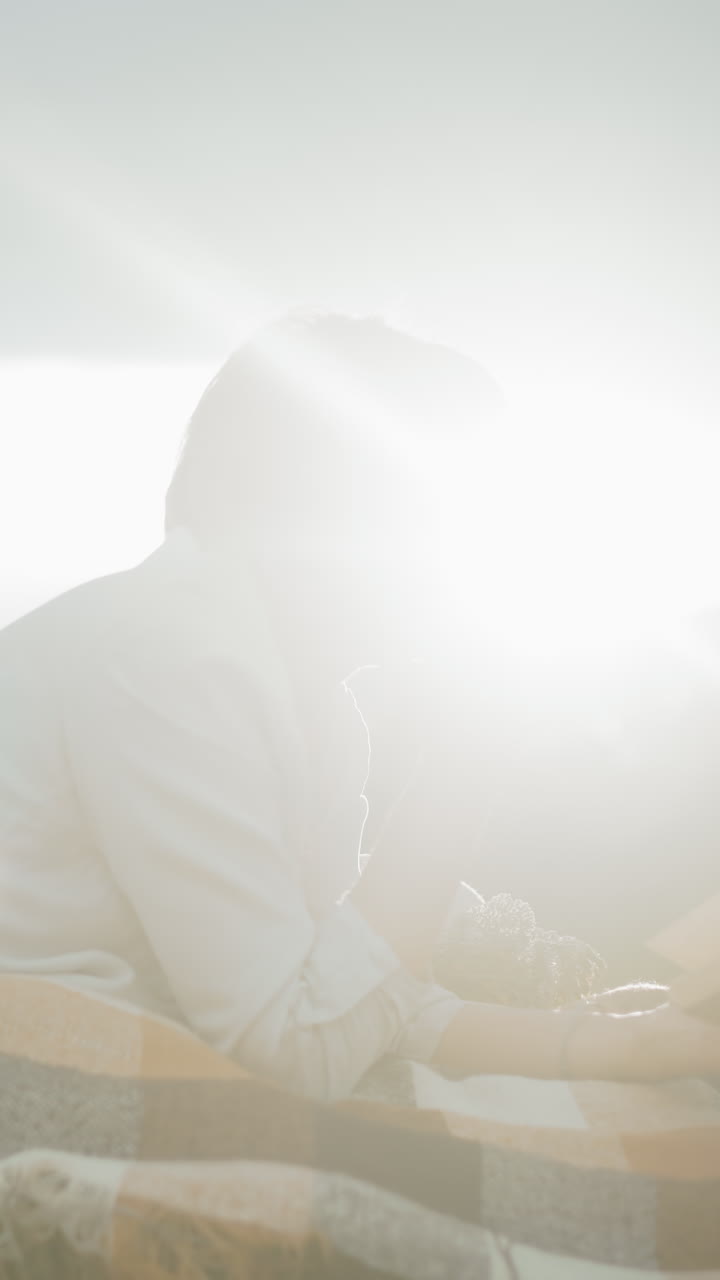 에 손을 고 책을 읽는 사려 깊은 여성, 광활한 야외에서  에 누워, 체크 된 담요는 따뜻한 자연 빛 아래 먼 지평선에 나무가 줄지어있는 동안 편안함을 더합니다.