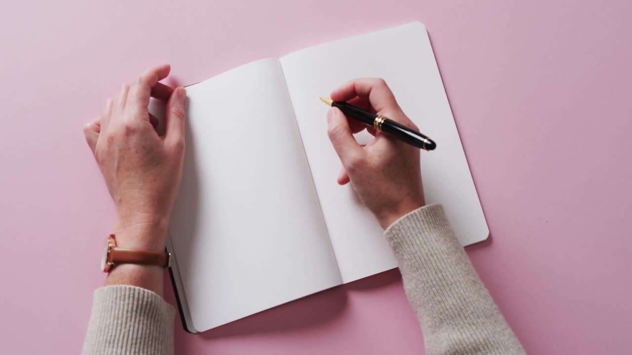 Close up of hand writing with pen on book with copy space on pink background in slow motion
