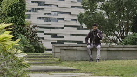 hombre caucásico bien vestido hablando por teléfono sentado en un banco de una zona residencial de la ciudad inteligente con edificios en el fondo en el área de parque verde con árboles y vegetación