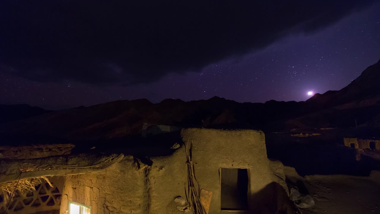 Vecchia casa di paese di notte con luna, stelle e la Nebulosa di Orione che si staglia sulle montagne in un paesaggio di sfondo.