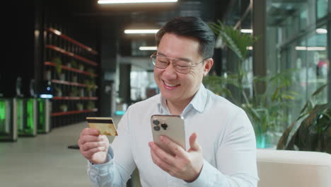 happy businessman using credit card and smartphone for online shopping payments in office lobby