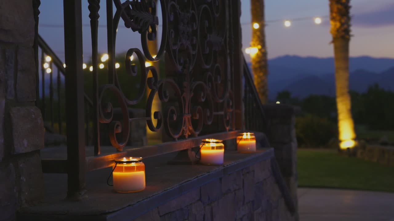 Romantic small flames from the candles on the side of a stone staircase with a string of lights in the background among the palm trees with a Revealing bonfire during sunset. Close up panning shot