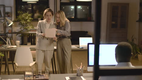 american man working with computer sitting at desk and two female coworkers are talking and reading a document