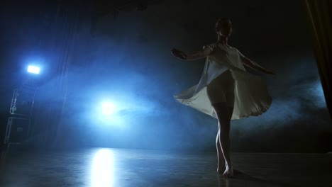 modern dance woman in a white dress dances a modern ballet, jumps, makes rotation on the stage with smoke in the blue spotlights.