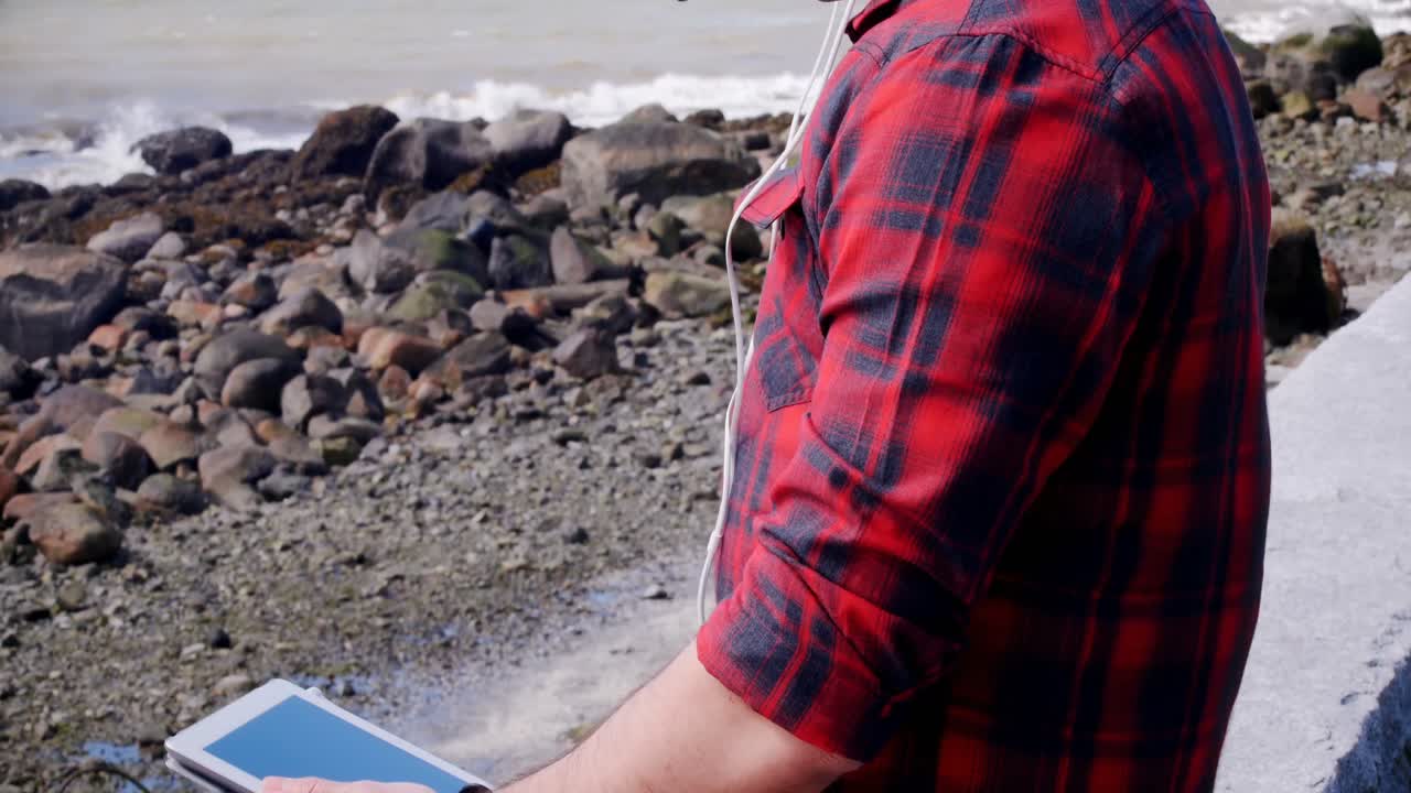 hombre con auriculares usando tableta digital en la playa 4k