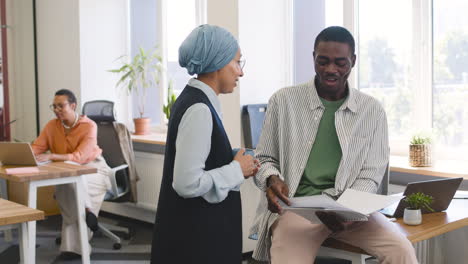 muslim businesswoman talks to a young worker who is holding a notebook and is leaning on his desk 1