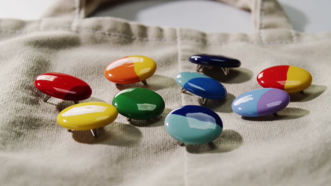 Bevegelig kamerafokus, 9 blanke emaljepins som viser glans på en beige handlenett, metallstifter synlige.