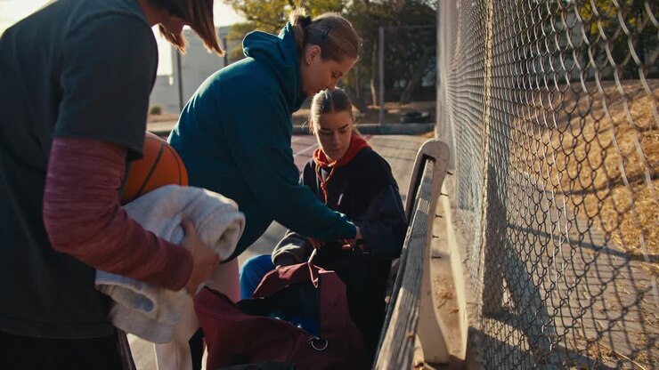 Three blonde girls in sportswear put their things in sports bags after training on an outdoor basketball court on a bench near a lattice fence on a sunny summer day